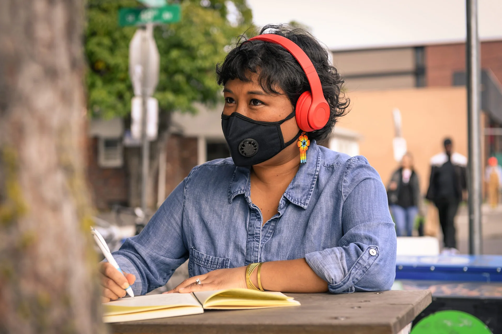 Person wearing headphones and a face mask writing in a notebook at an outdoor table.