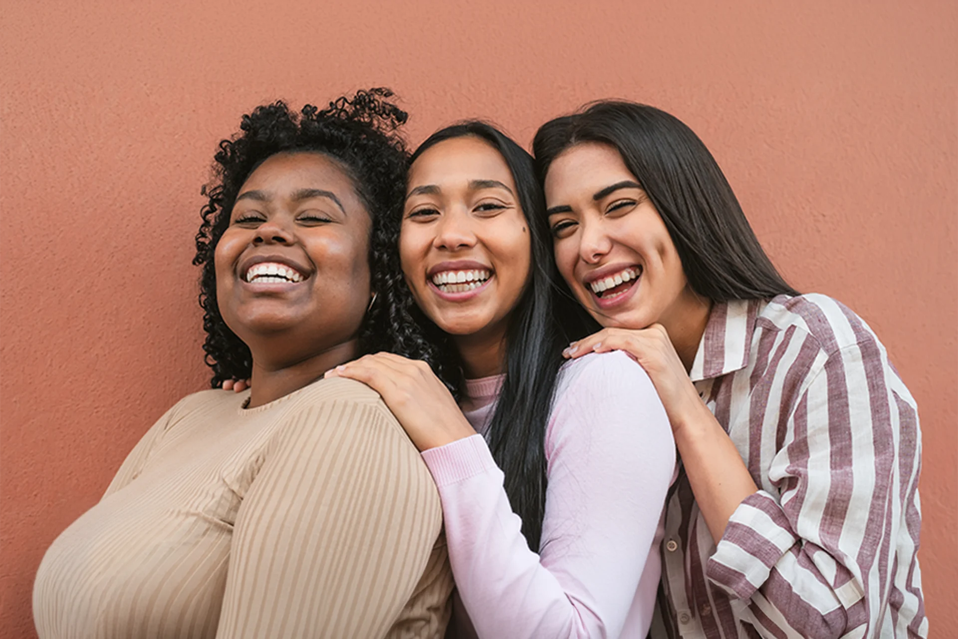 Three diverse young people smiling and laughing together against a warm-colored wall.