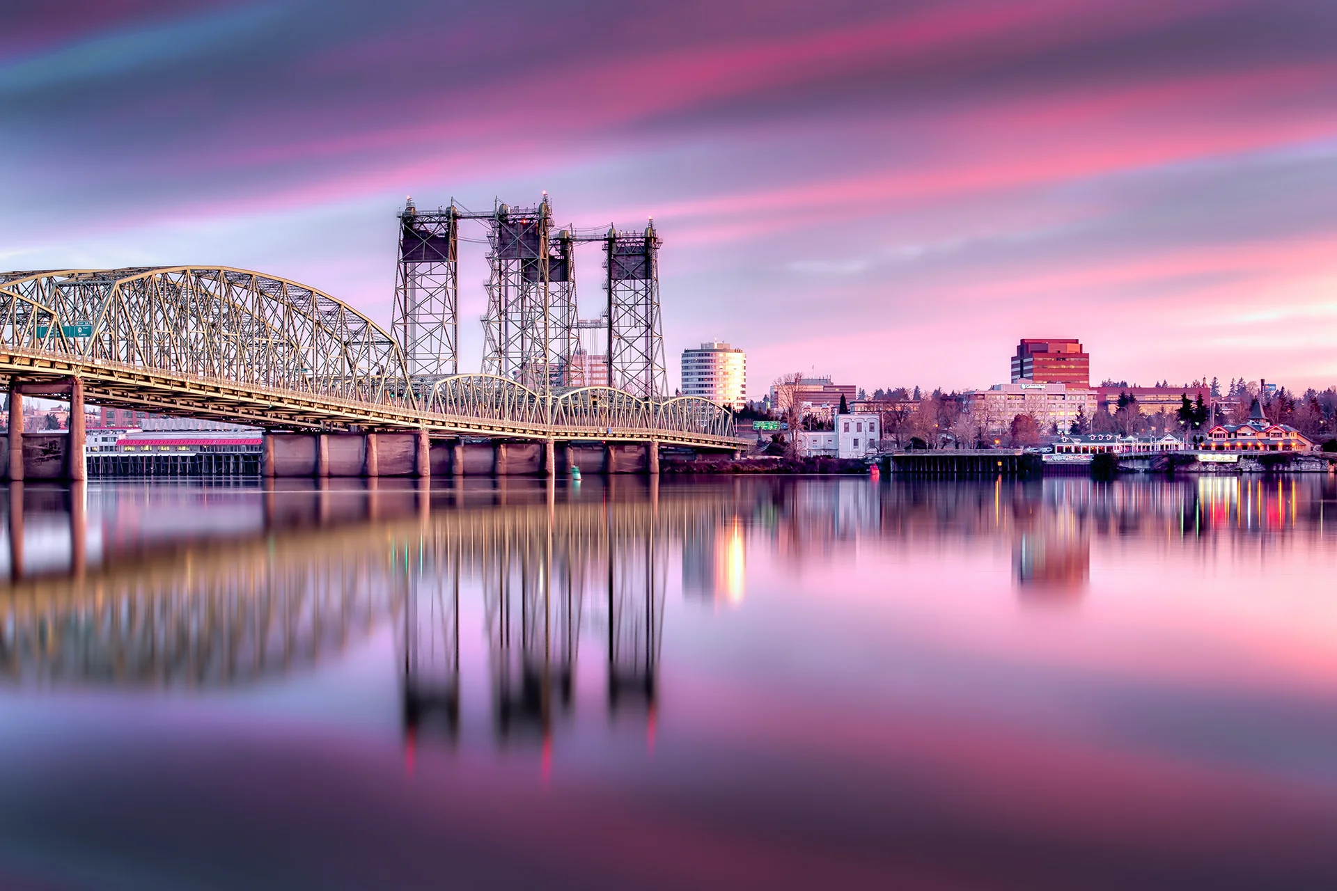 Interstate Bridge crossing the Columbia River beside the Vancouver, Washington waterfront under a colorful sunset sky.