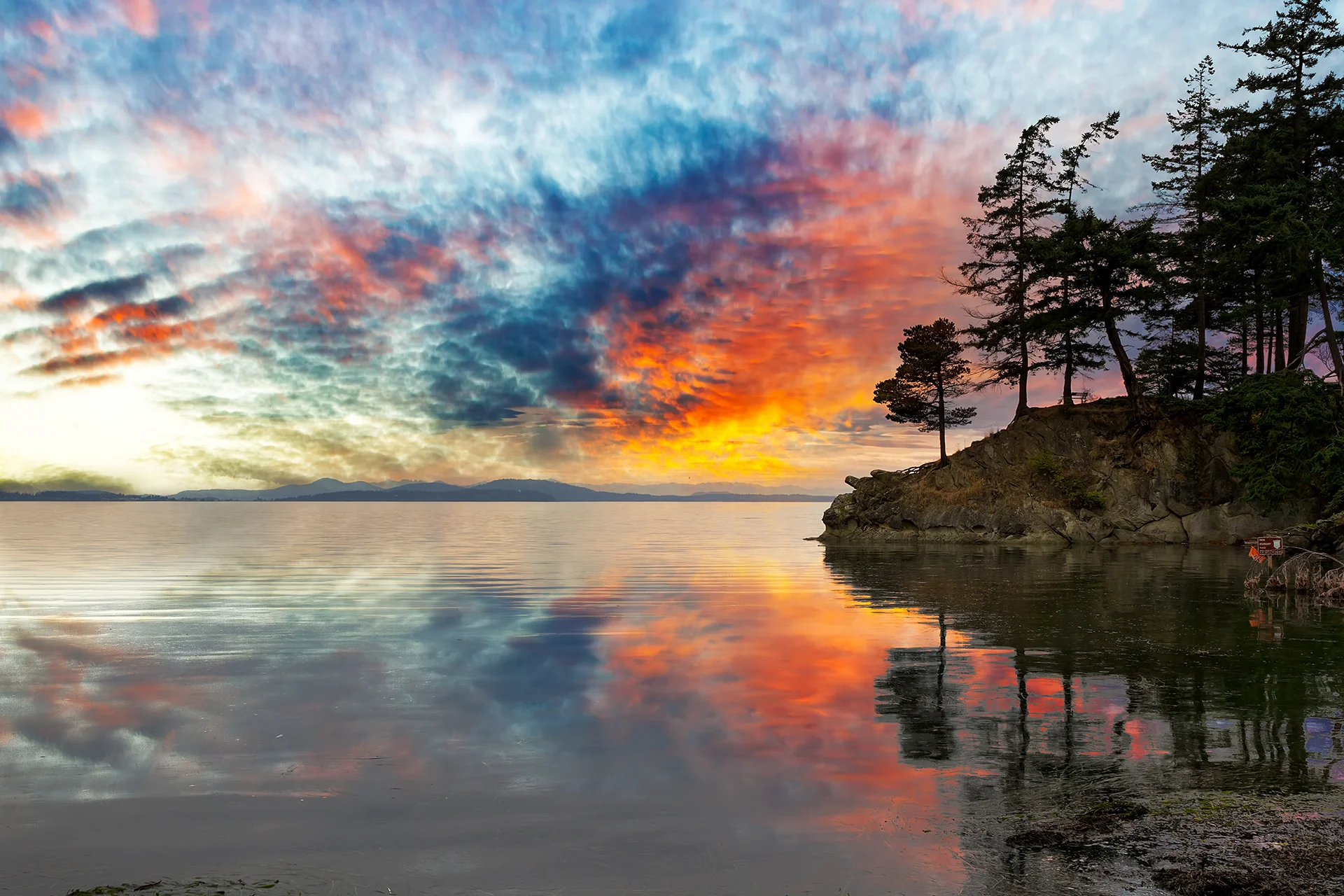 Colorful sunset reflected across calm Washington waters beside a rocky shoreline lined with evergreen trees.